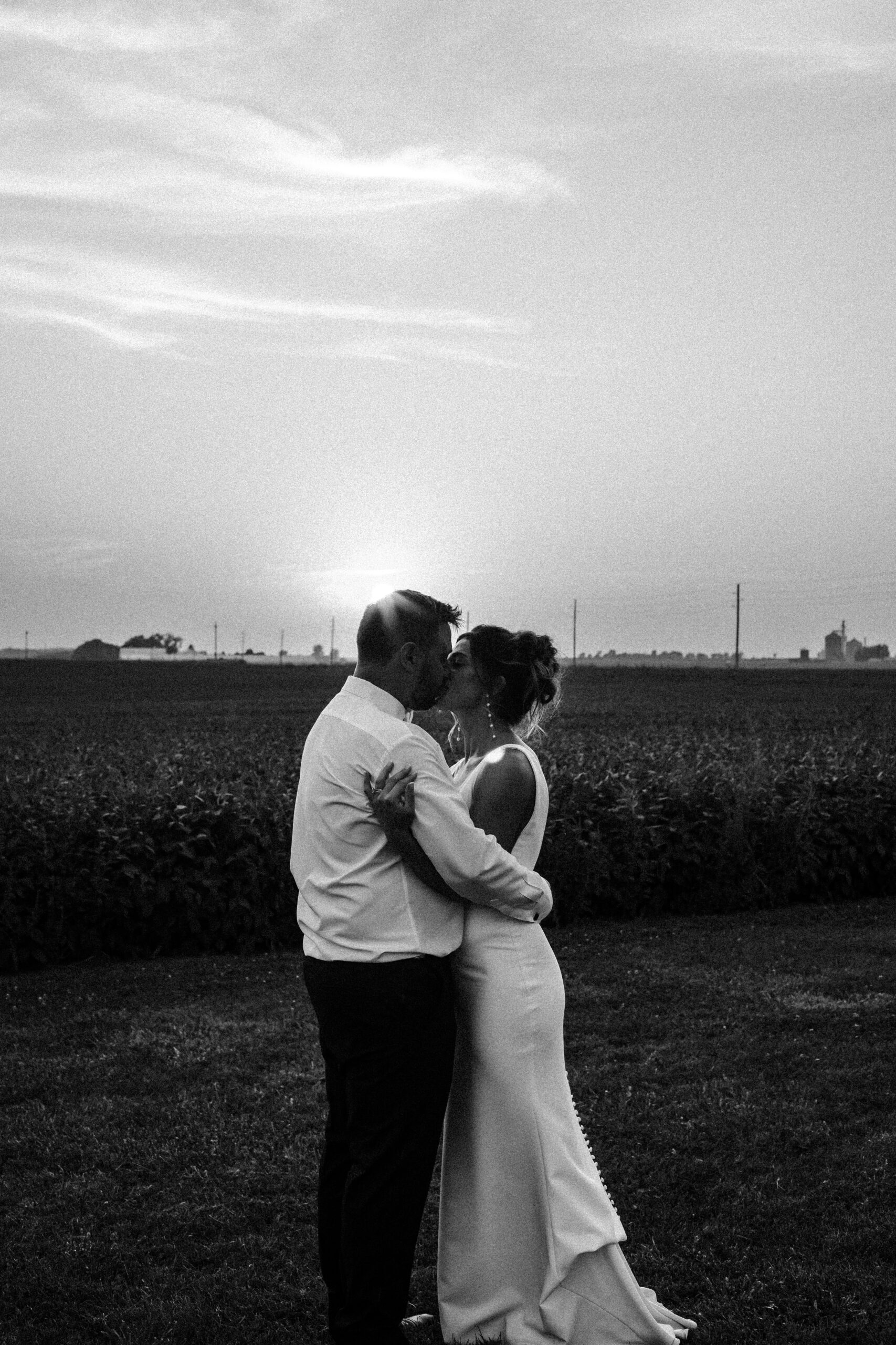 Bride and groom share a kiss at sunset in a field, silhouetted against the sky.